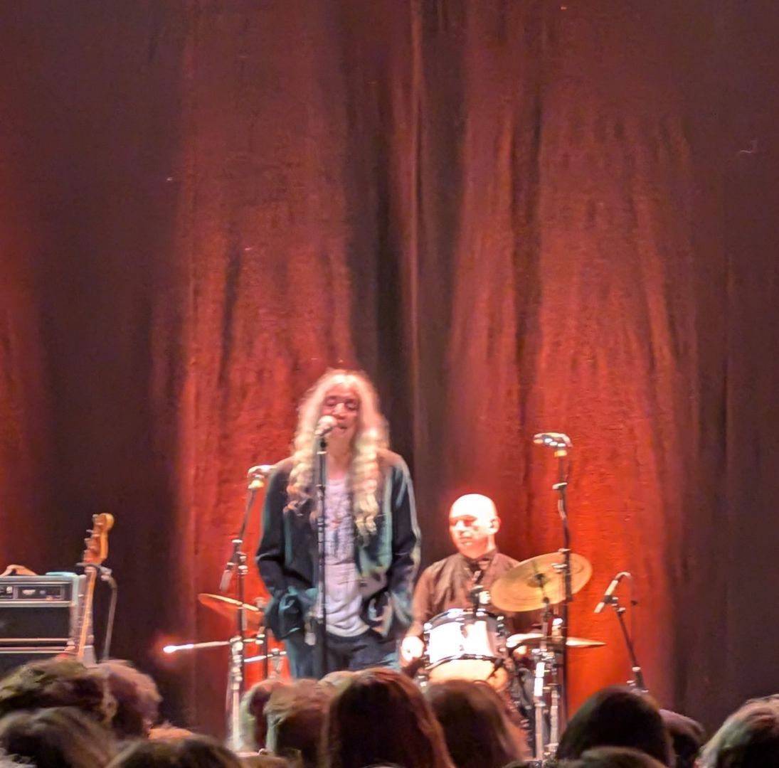 A slightly blurry photo of Patti Smith on stage. A red curtain forms the backdrop, lit from below. Behind her is the drummer. Patti herself has long white hair and stands casually singing into the microphone with her hands in her pockets.