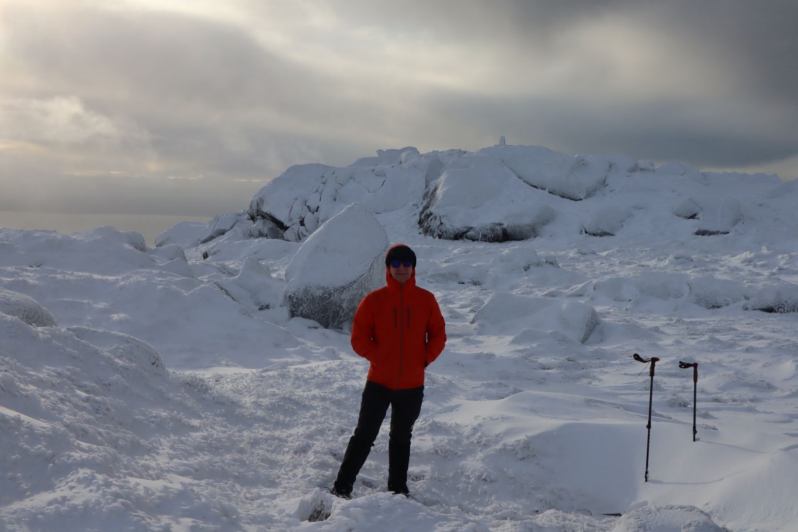 A man, me, on a snowy hilltop. There are also a pair of walking poles stuck in the snow.