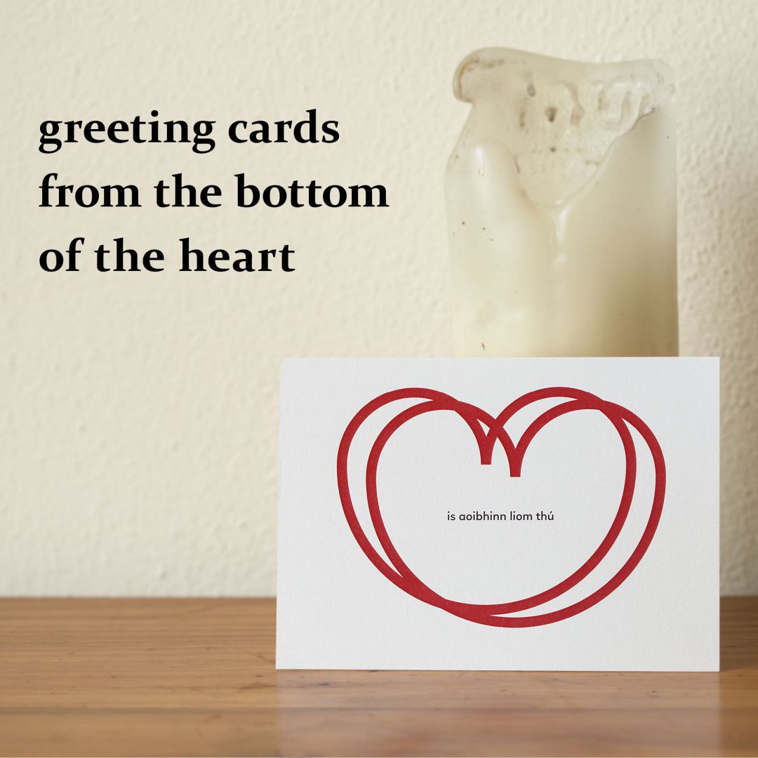 the image shows a photogrtaph of a love card standing on a wooden desk, leaning against a white candle in front of a cream coloured wall. in the top left corner is the headline 'greeting cards from the bottom of the heart' in black bold lower case letters. the greeting card shows the irish sentence ‚is aoibhinn liom thú‘, which means 'i love you' within two overlapping red heart outlines.