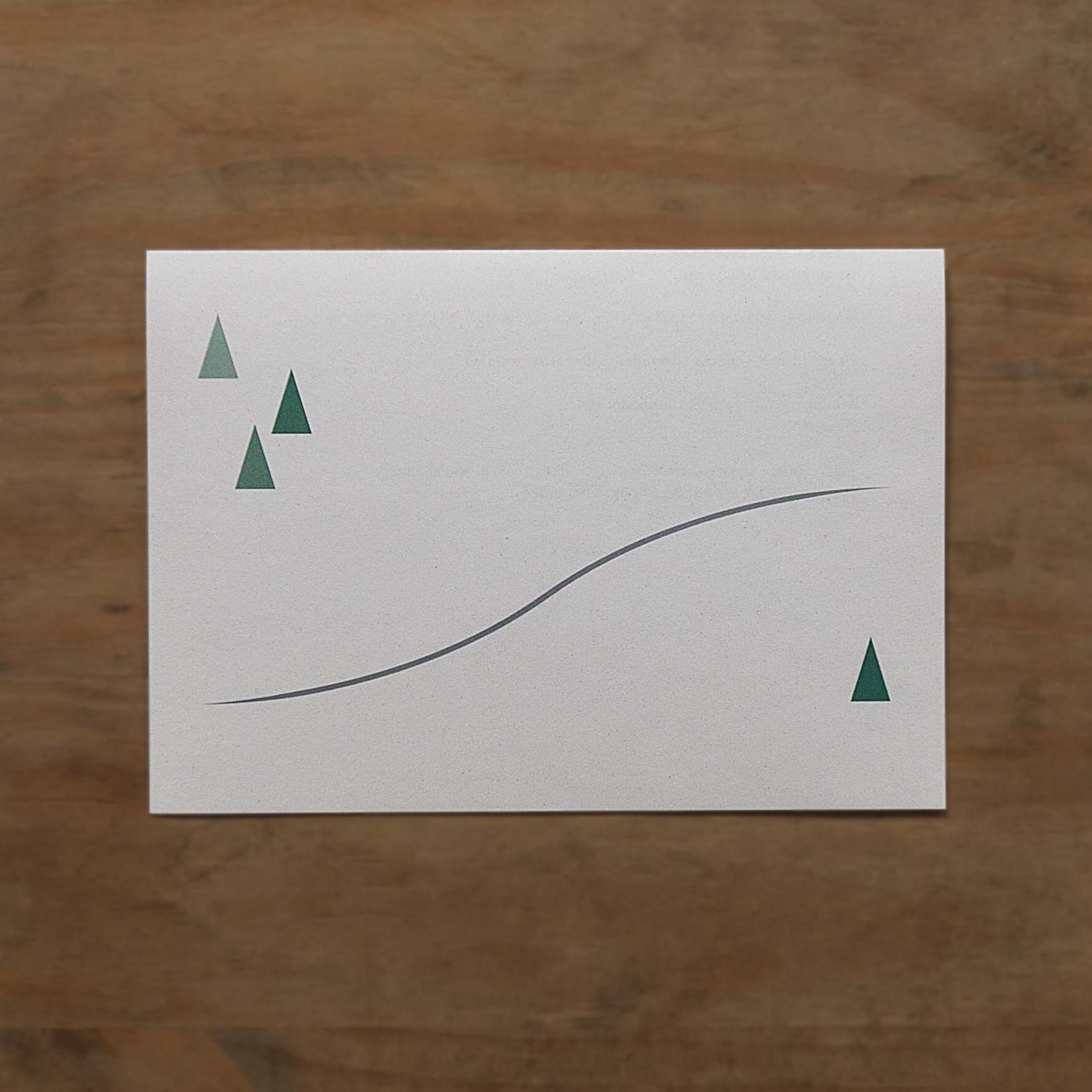 the photograph shows the back of a folded letter, which shows three green triangles in the top left vorner illustrating trees, and one in the bottom left corner. inbetween is a grey meandering path. the unprinted ares illustrates snow.