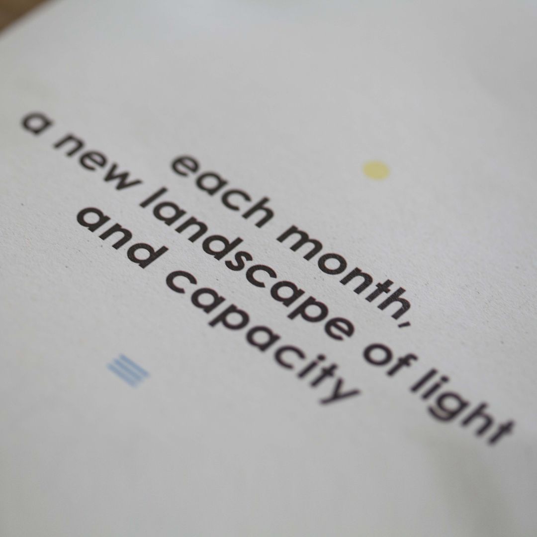the image shows a photograph of a headline of a front of a brochure on a wooden desk in an angle. the headline says 'each month, a new landscape of light and capacity' in black bold lower case letters running over three rows. above the headline is a yellow corcle and underneath thre blue lines.